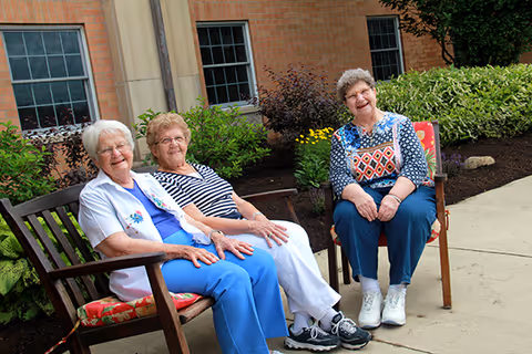 Three elderly women sitting outside on chairs near a building with brick walls and windows, smiling and enjoying the outdoor setting with greenery and flowers behind them.