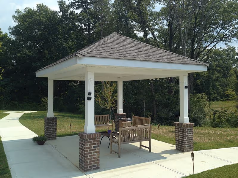 A small outdoor pavilion with a shingled roof supported by four white columns with brick bases. Under the pavilion are wooden benches and a small table. The pavilion is situated on a concrete path surrounded by grass and trees in the background.