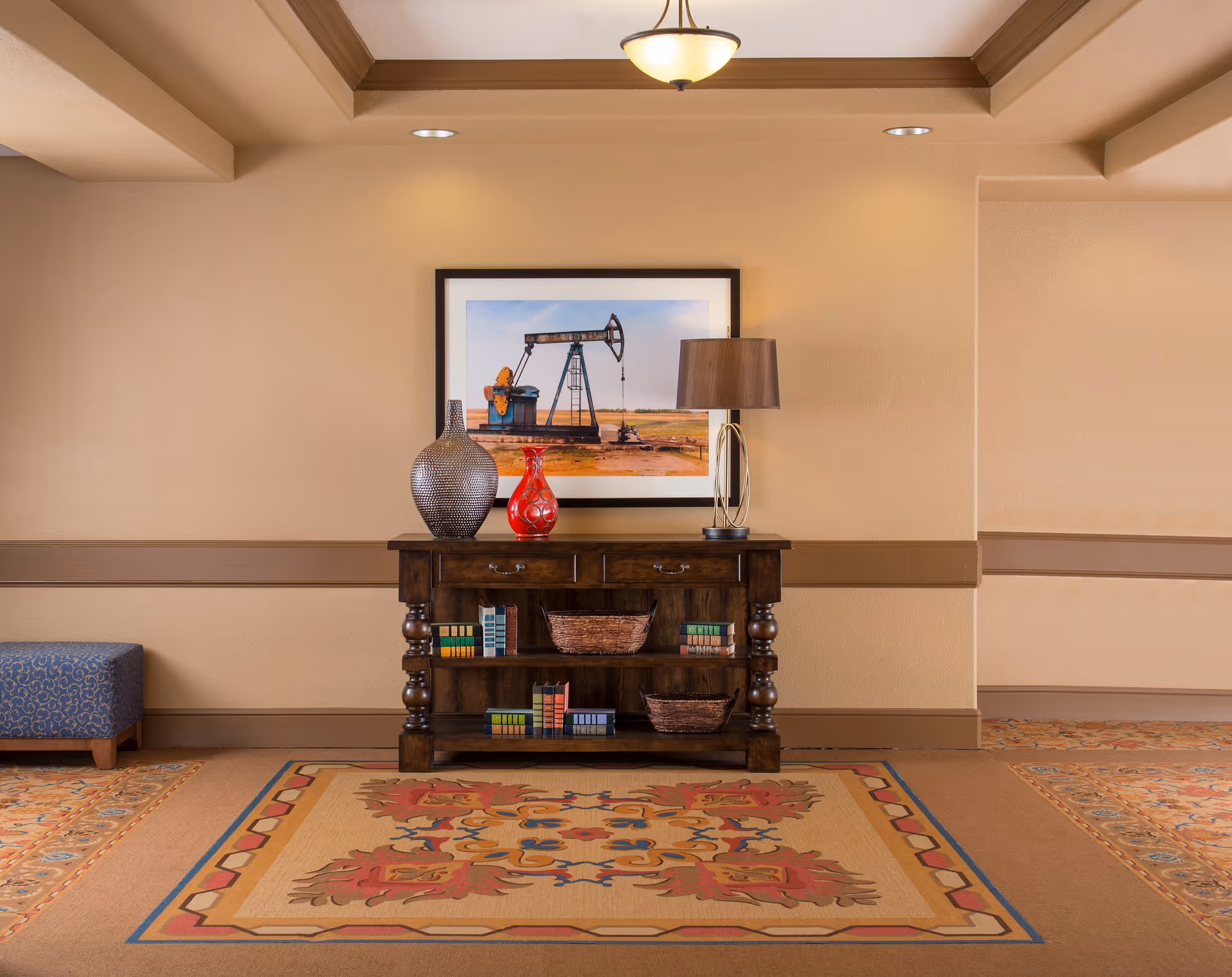 A decorative wooden console table with two drawers and shelves holding books and baskets, topped with a large textured vase, a smaller red vase, and a modern table lamp. Above the table is a framed photograph of an oil pumpjack in a field. The room has beige walls with brown trim, a ceiling light fixture, and a patterned carpet with warm colors.