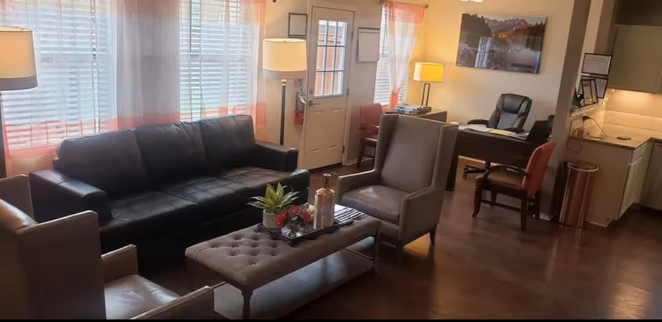 A cozy living room area with a black leather sofa, two gray armchairs, and a tufted ottoman coffee table with decorative items on it. There are two floor lamps near large windows with sheer white and orange curtains. In the background, there is a desk with office chairs, a wall-mounted picture, and a small kitchen area with cabinets and a trash can.