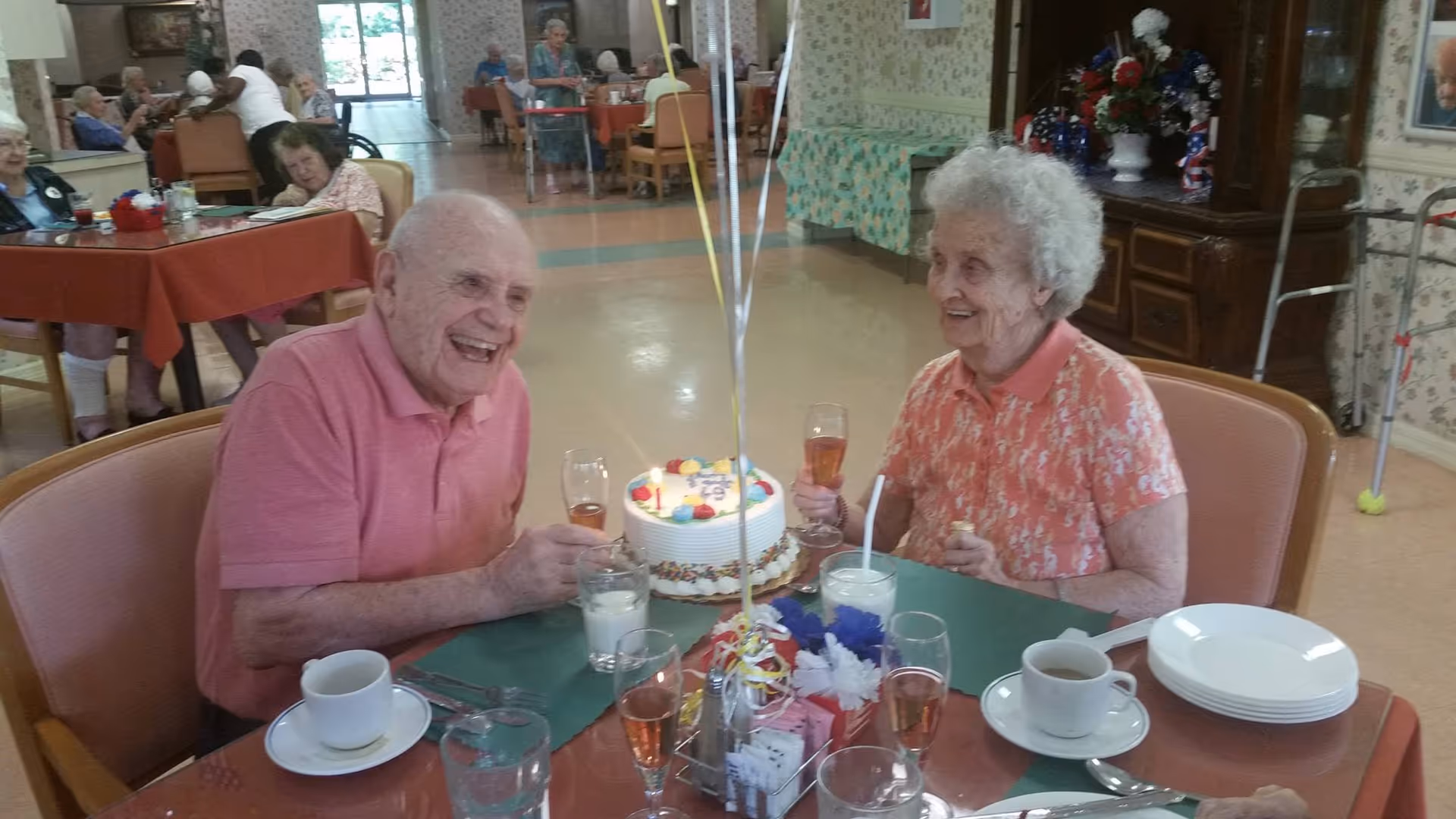 Two elderly people sitting at a dining table in a senior living facility, smiling and holding glasses of a pink beverage. A decorated birthday cake with candles and balloons is on the table between them. Other residents are visible in the background seated at tables.