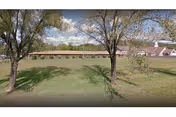 Wide outdoor view of a grassy lawn, two trees, and a long building in distance.