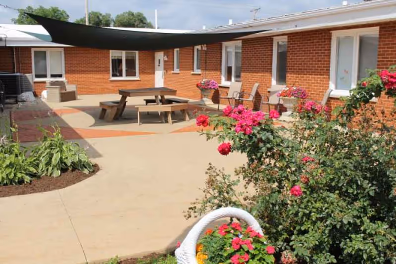 Outdoor courtyard area of a senior living facility with brick walls, several windows, and doors. The courtyard features a shaded seating area with a picnic table and benches, potted flowers, and landscaped flower beds with blooming pink and red flowers.