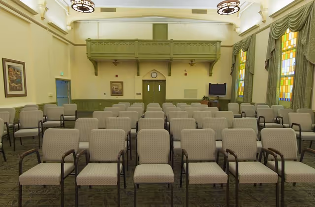 An empty auditorium-style room with rows of upholstered chairs facing double doors, stained glass windows, and a small balcony.