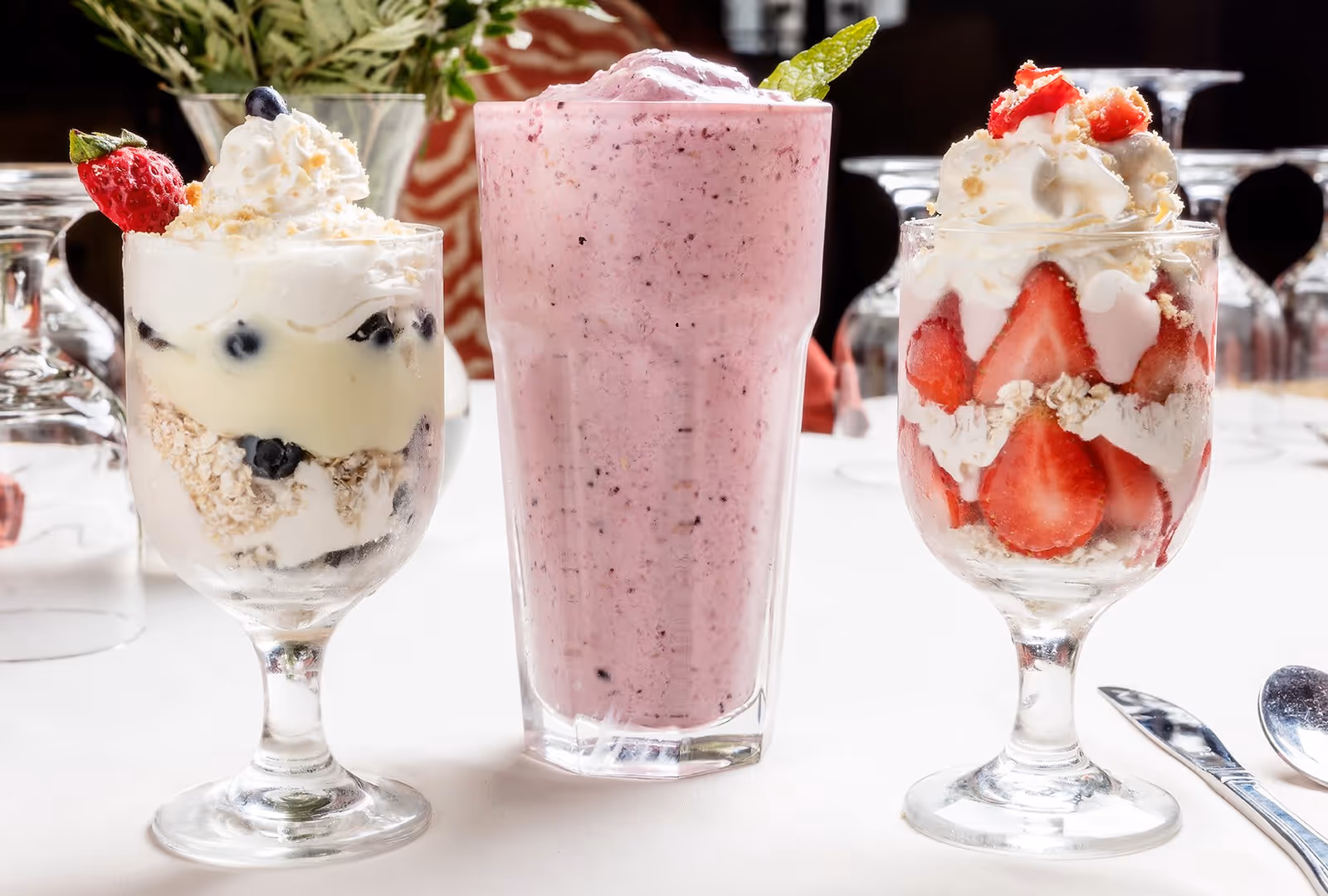 Three dessert glasses on a white tablecloth. The left glass contains a layered dessert with blueberries, cream, and a strawberry garnish. The middle glass holds a pink smoothie topped with a mint leaf. The right glass has a layered dessert with sliced strawberries, cream, and whipped cream topping.