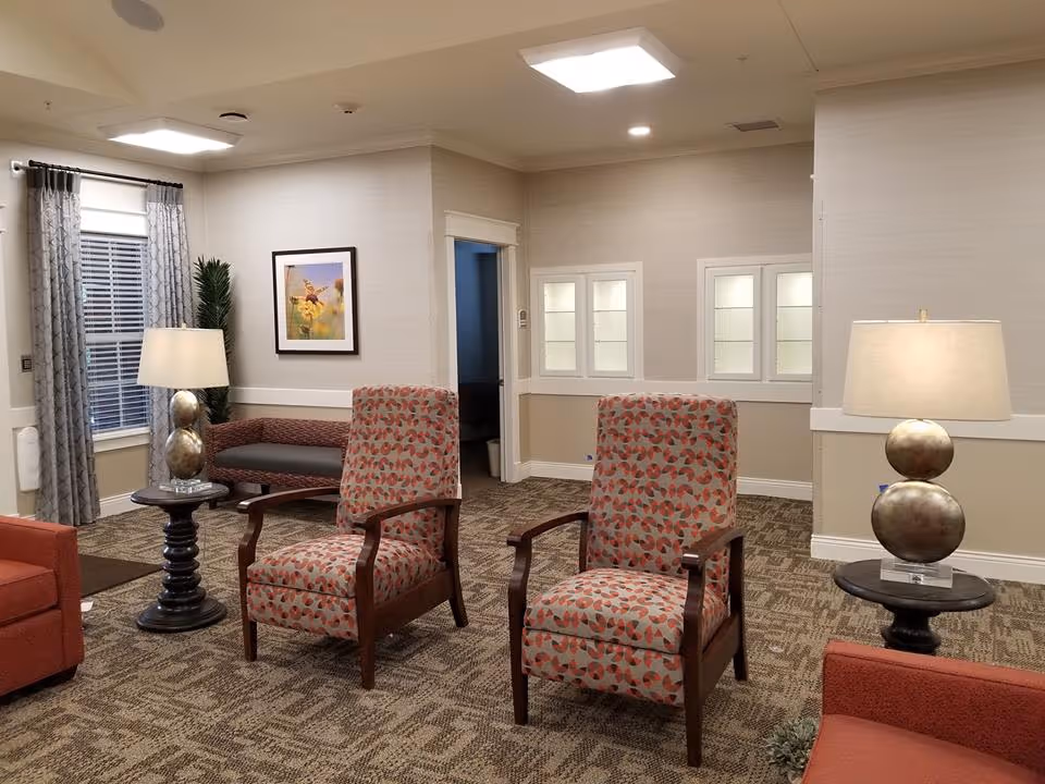 A cozy living room area with two patterned armchairs, a red sofa, two round side tables each with a lamp, a window with curtains, a framed picture on the wall, and a bench with a plant beside it.