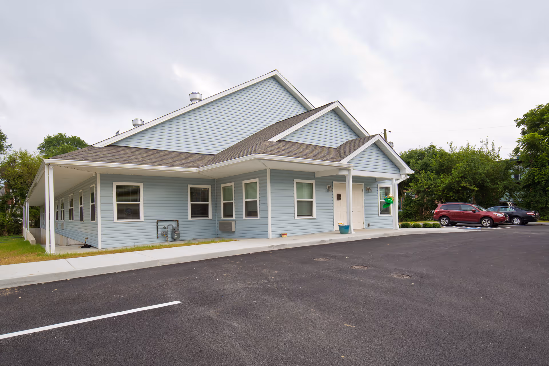 Exterior view of a light blue single-story building with white trim, a covered entrance with two white doors, and a parking lot with a few parked cars. The sky is cloudy and there are trees and greenery around the building.