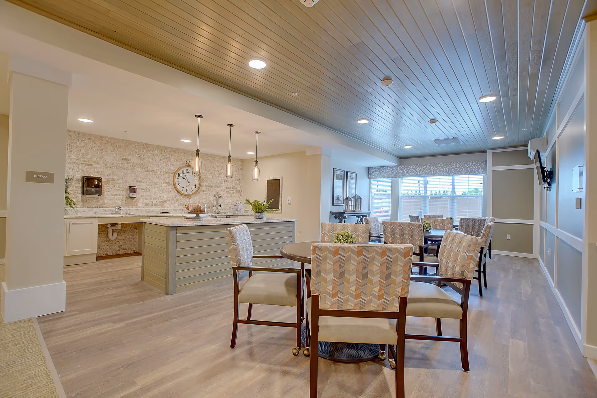Bright communal dining area with round tables and patterned chairs facing a bistro-style counter and wood-paneled ceiling.