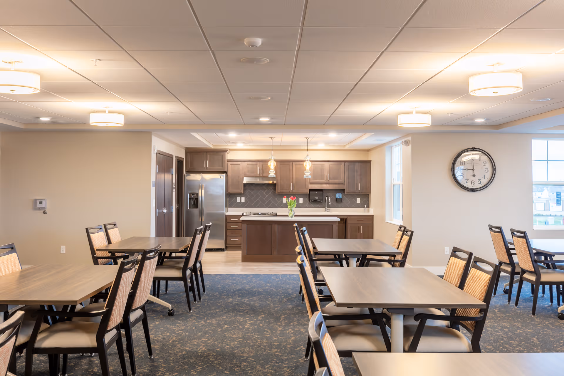 A bright communal dining room with multiple tables and chairs facing a kitchen area with a central island, stainless steel refrigerator, and wall clock.