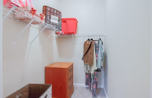 A small walk-in closet with a wooden chest of drawers, a few hanging clothes on a white wire rack, and storage bins on the upper shelf. The walls are light-colored and the floor is carpeted.