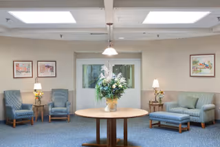 A bright seating lounge with upholstered chairs and a sofa arranged around a central round table holding a large floral arrangement.
