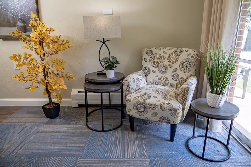 A cozy corner in a senior living facility featuring a floral-patterned armchair, two round nesting tables with a small potted plant and a lamp, a side table with a tall green plant in a white pot, and a decorative yellow leafy plant in a black pot. The area has a blue and gray patterned carpet and a large window with beige curtains letting in natural light.