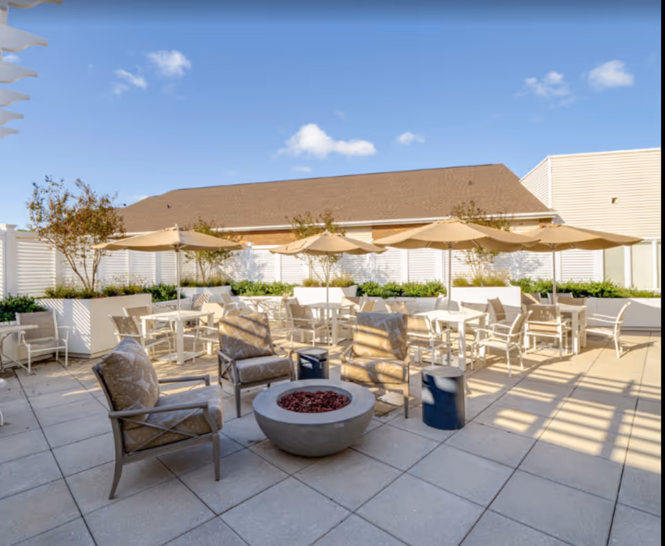Sunlit outdoor patio with chairs, tables, umbrellas and a central fire pit.
