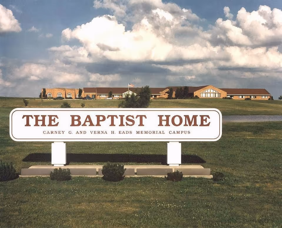 Wide view of a lawn with a sign reading "The Baptist Home" and the facility building under a cloudy sky in the background.
