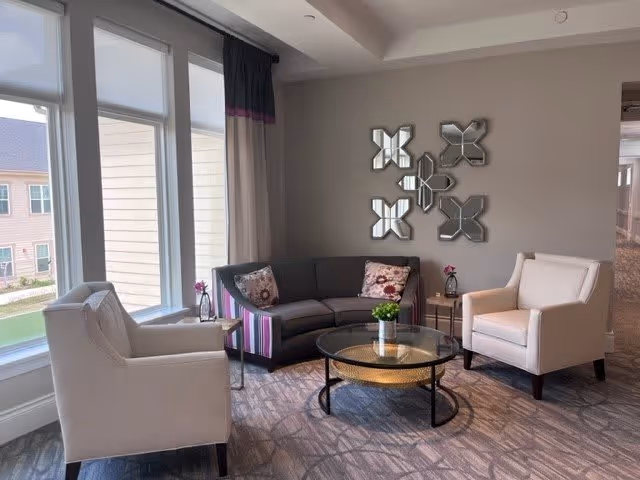 A cozy living room area with a gray curved sofa adorned with floral pillows, two white armchairs, a round glass coffee table with a small green plant on it, and a decorative wall mirror featuring multiple geometric shapes. Large windows with white blinds and curtains allow natural light to fill the room.