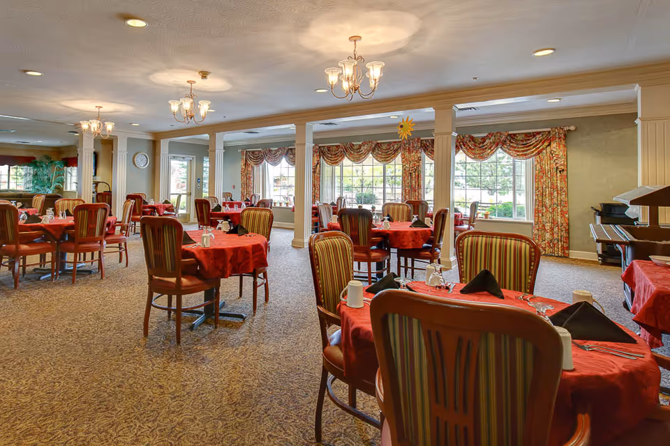 Spacious dining room with round tables dressed in red tablecloths and striped chairs beneath chandeliers and large windows.