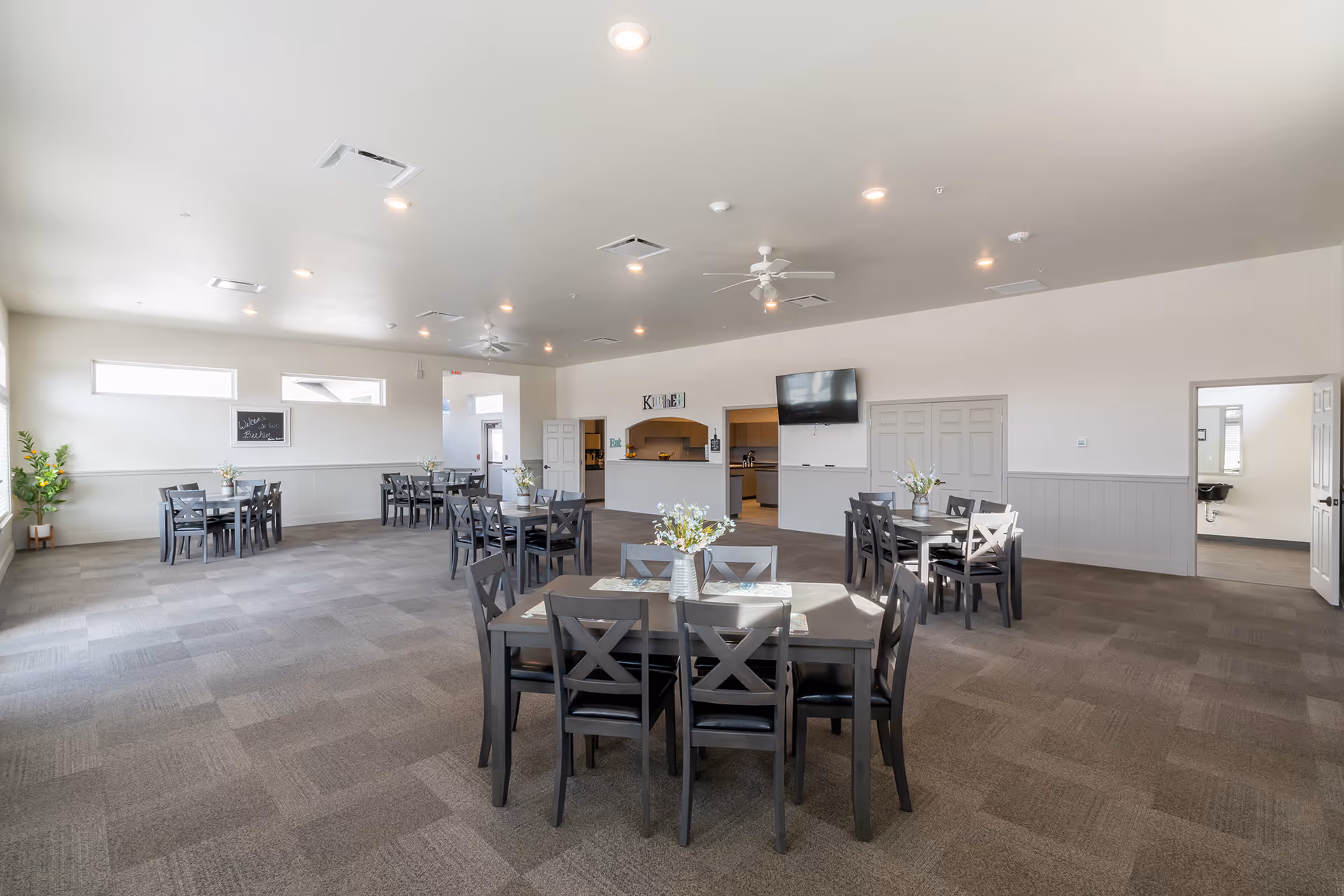 A spacious dining room with multiple dark wooden tables and chairs arranged neatly. Each table has a small floral centerpiece. The room has light-colored walls, carpeted flooring, ceiling fans, and recessed lighting. There is a kitchen area visible in the background with a sign above it that says 'KITCHEN'. A flat-screen TV is mounted on the wall near the kitchen. There are windows letting in natural light and a door leading to another room.