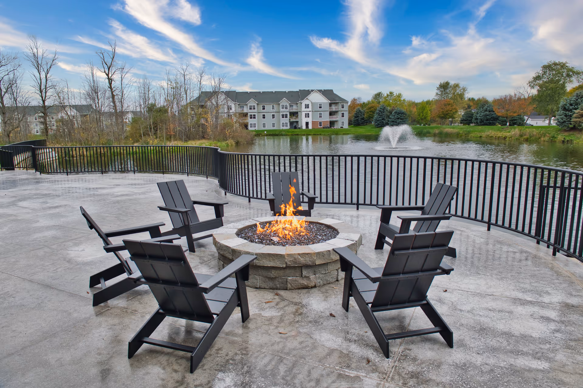 Outdoor patio area with six black Adirondack chairs arranged around a stone fire pit with a fire burning. The patio overlooks a pond with a water fountain, surrounded by trees and a multi-story residential building in the background under a partly cloudy sky.