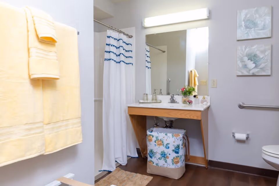 A clean and well-lit bathroom with a wooden vanity and white countertop. There is a large mirror above the sink with a horizontal light fixture. A floral-patterned laundry basket is placed under the sink. To the left, a shower area with a white curtain featuring blue accents is visible. Yellow towels hang on a rack on the left wall. The bathroom has light gray walls, two floral wall decorations, a toilet paper holder, and a grab bar next to the toilet.