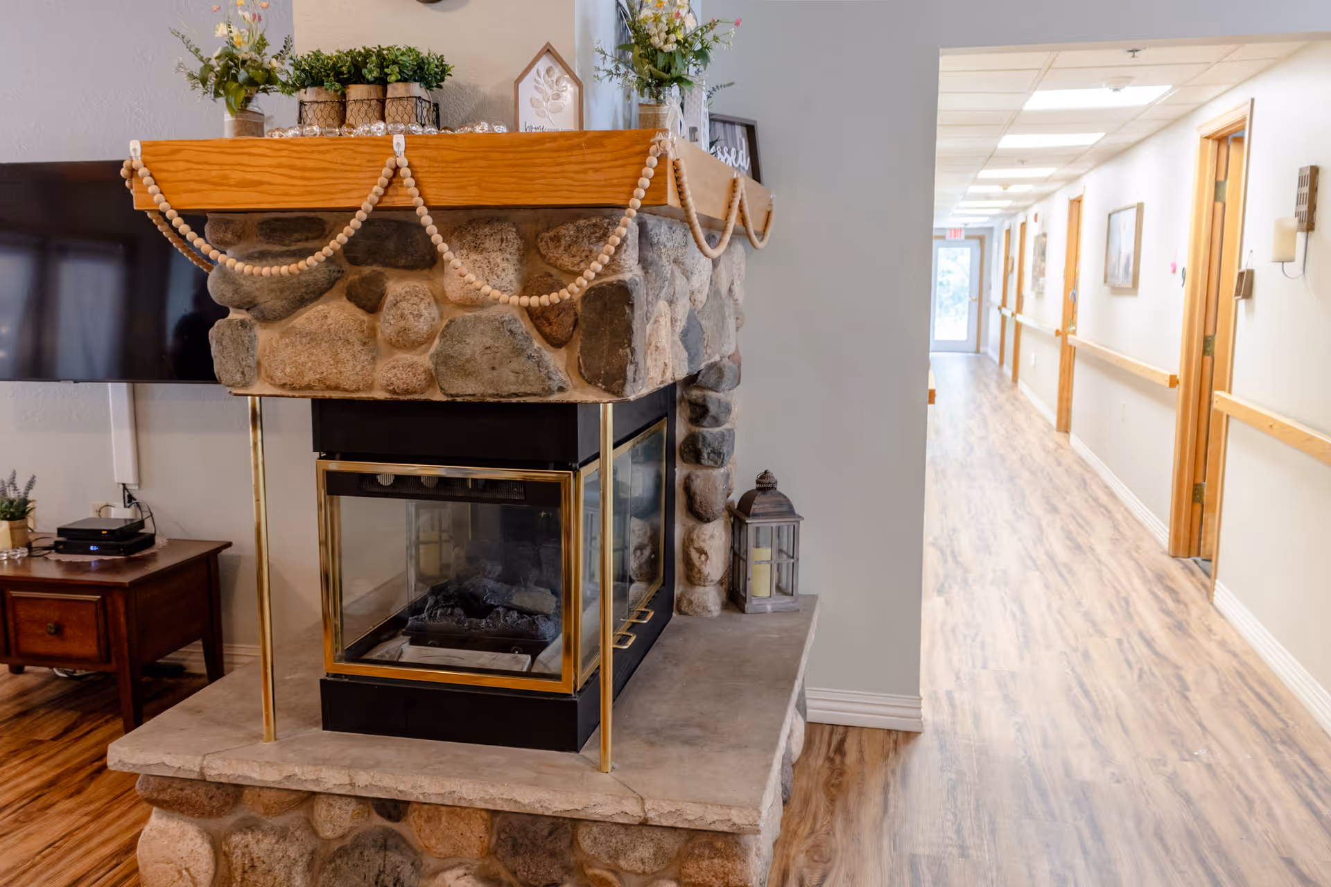 Interior view of a senior living facility hallway with wooden flooring and light-colored walls. On the left side, there is a stone fireplace with a wooden mantle decorated with plants and ornaments. A flat-screen TV and a wooden side table with a small plant and electronic device are also visible. The hallway has several doors and a handrail along the wall, leading to a glass exit door at the far end.