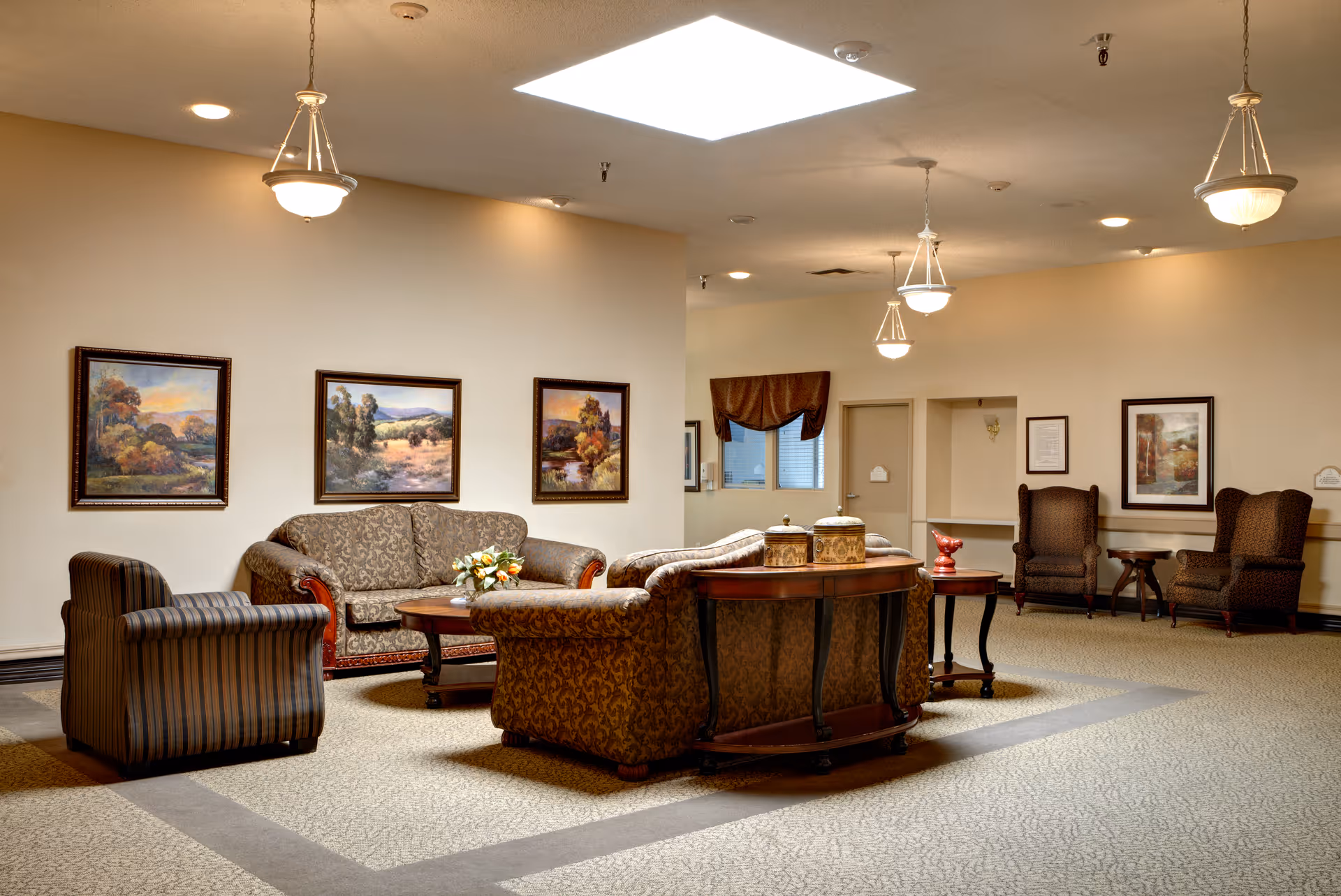 A spacious and well-lit living room area in a senior living facility with patterned sofas and armchairs arranged around a wooden coffee table. The walls are decorated with framed landscape paintings, and the ceiling has multiple hanging light fixtures and a skylight. There are additional chairs and small tables along the walls, creating a comfortable and inviting common area.
