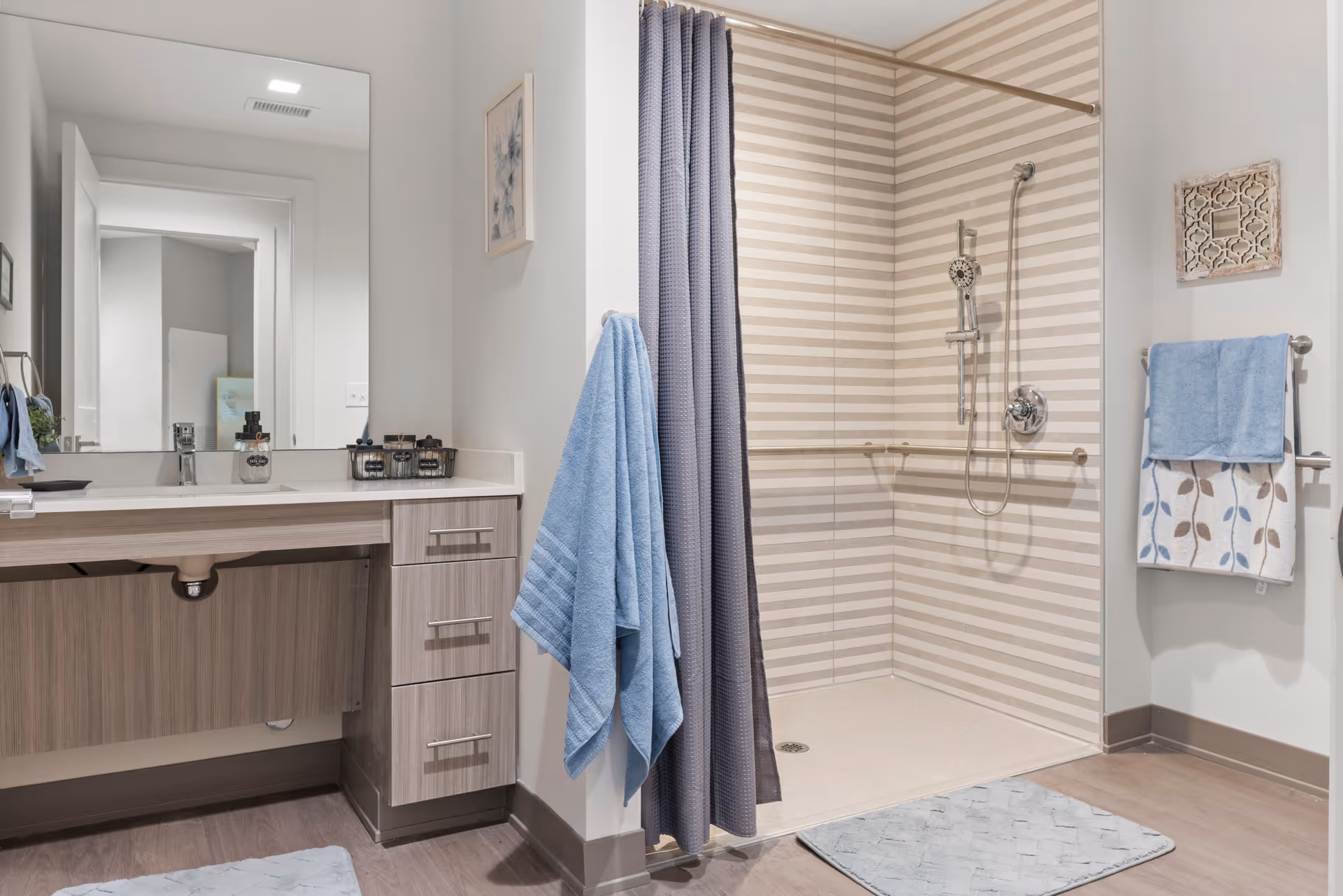 A modern bathroom featuring a walk-in shower with beige and white horizontal striped tiles, a handheld showerhead, and grab bars. There is a blue shower curtain and matching blue towels hanging on the wall. A vanity with a large mirror, sink, and wooden drawers is visible on the left side. The floor has a light blue bath mat.