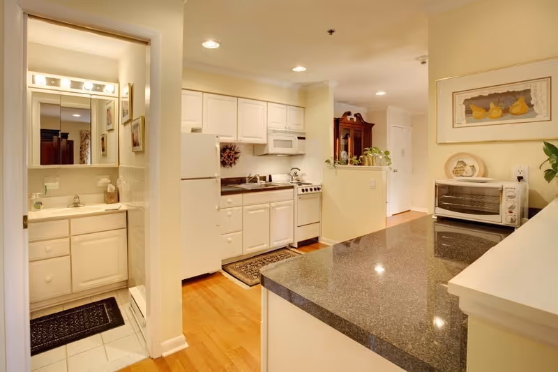 Small bright kitchen with white cabinets, a dark granite countertop island, appliances and a small sink/bathroom visible to the left.