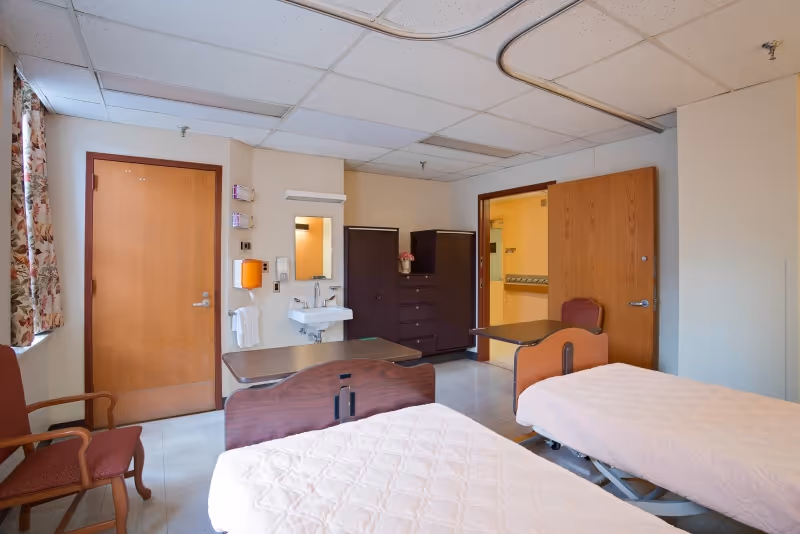 A room with two hospital-style beds covered with white quilts, a wooden chair, a small sink with a mirror above it, two wooden wardrobes, and two wooden doors. The room has a tiled floor and a drop ceiling with fluorescent lights. A window with floral curtains is partially visible on the left side.