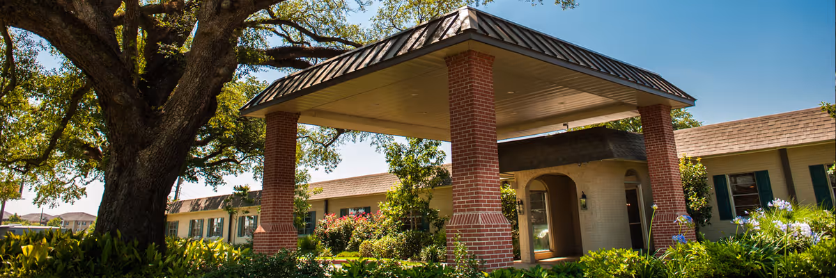 Exterior view of Lady of the Oaks Retirement facility showing a large covered entrance supported by brick pillars, surrounded by lush greenery and a large tree on the left side.