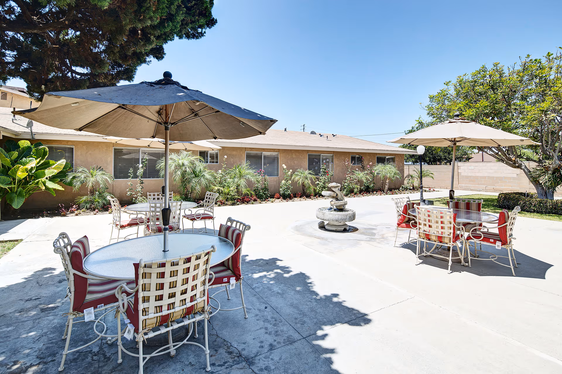 Outdoor courtyard area with two round tables, each surrounded by four chairs with red and white cushions. Each table has a large beige umbrella providing shade. There is a small decorative fountain in the center of the courtyard, surrounded by a concrete patio. The background shows a single-story building with windows and various green plants and trees around the perimeter under a clear blue sky.