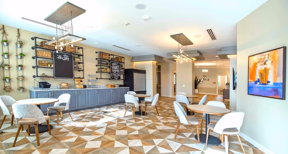 Bright, modern common dining/cafe area with round tables and upholstered chairs, a service counter with shelving, and geometric wood flooring.