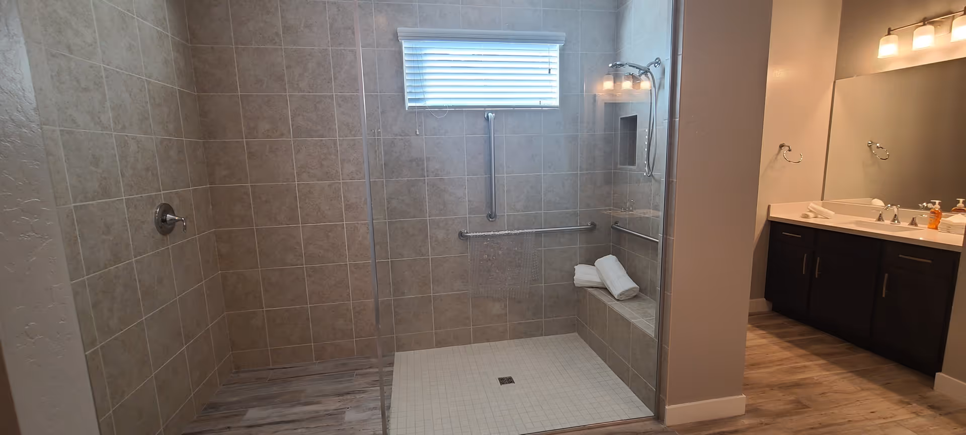 A modern bathroom featuring a walk-in shower with beige tiled walls and a glass partition. The shower has a built-in bench with two folded white towels, a handheld showerhead, and grab bars for safety. Next to the shower is a vanity with a large mirror, dark wood cabinets, and a countertop with two sinks and soap dispensers. The floor has a wood-like finish, and a small window with blinds is above the shower.