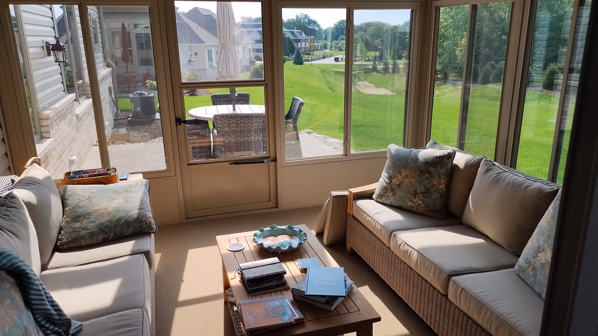 A sunlit enclosed patio area with two cushioned wicker sofas facing each other and a wooden coffee table in between. The coffee table holds several books and a decorative bowl. Large windows and a glass door provide a view of a green lawn and outdoor patio furniture with an umbrella outside.