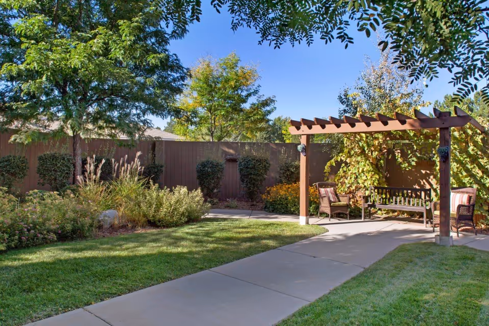 A peaceful outdoor garden area with a paved walkway, green grass, various bushes and trees, and a wooden pergola with a bench and two cushioned chairs underneath. The sky is clear and blue.