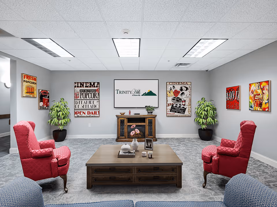 A cozy lounge area with two red upholstered armchairs facing a wooden coffee table decorated with vases and candles. The walls are adorned with colorful vintage-style movie posters and two potted plants are placed on either side of a wooden cabinet with a Trinity Hills of Knoxville sign above it. The room has a gray carpet and ceiling lights.