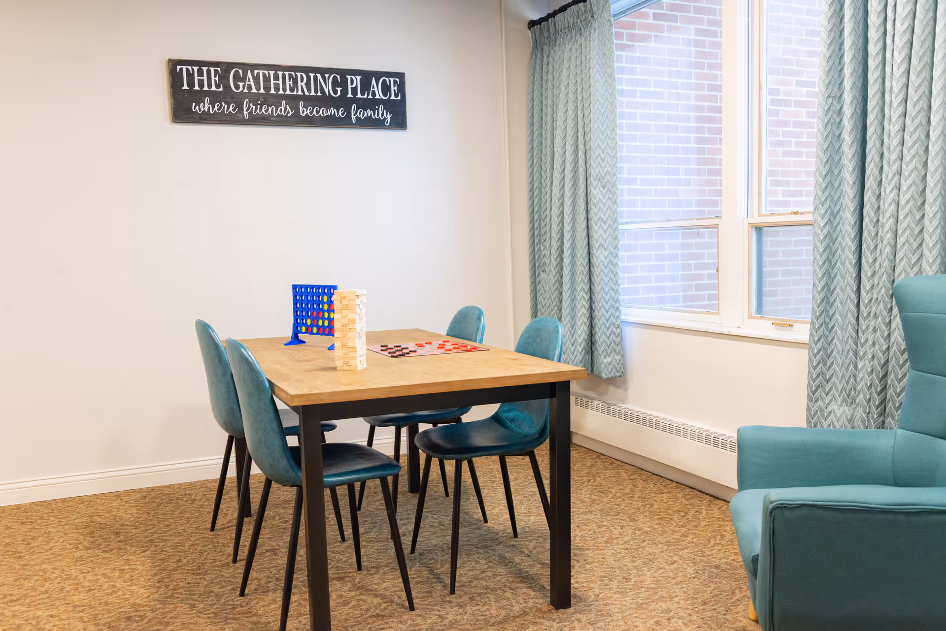 A bright communal room with a wooden table and teal chairs arranged with board games under a wall sign reading "The Gathering Place".