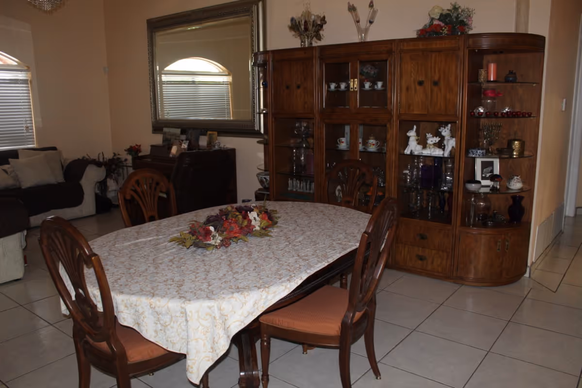 A dining room with a table covered by a patterned tablecloth and floral centerpiece, surrounded by wooden chairs, with a large wooden china cabinet and a living area visible in the background.