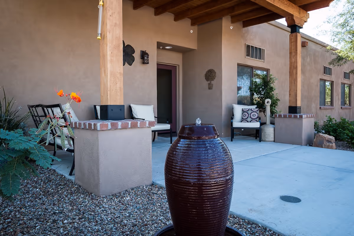 Covered stucco patio with wooden posts, cushioned chairs and a large ceramic fountain urn in the foreground.