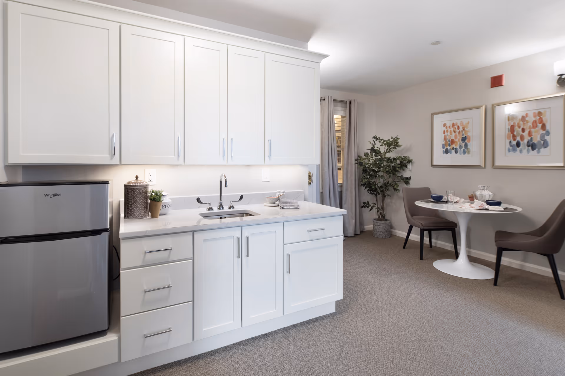 A clean and modern kitchenette area with white cabinets, a small stainless steel Whirlpool refrigerator, and a sink with a chrome faucet. To the right, there is a small round white dining table set with two gray chairs, some tableware, and two framed abstract paintings on the wall. A potted plant is placed near a window with gray curtains.