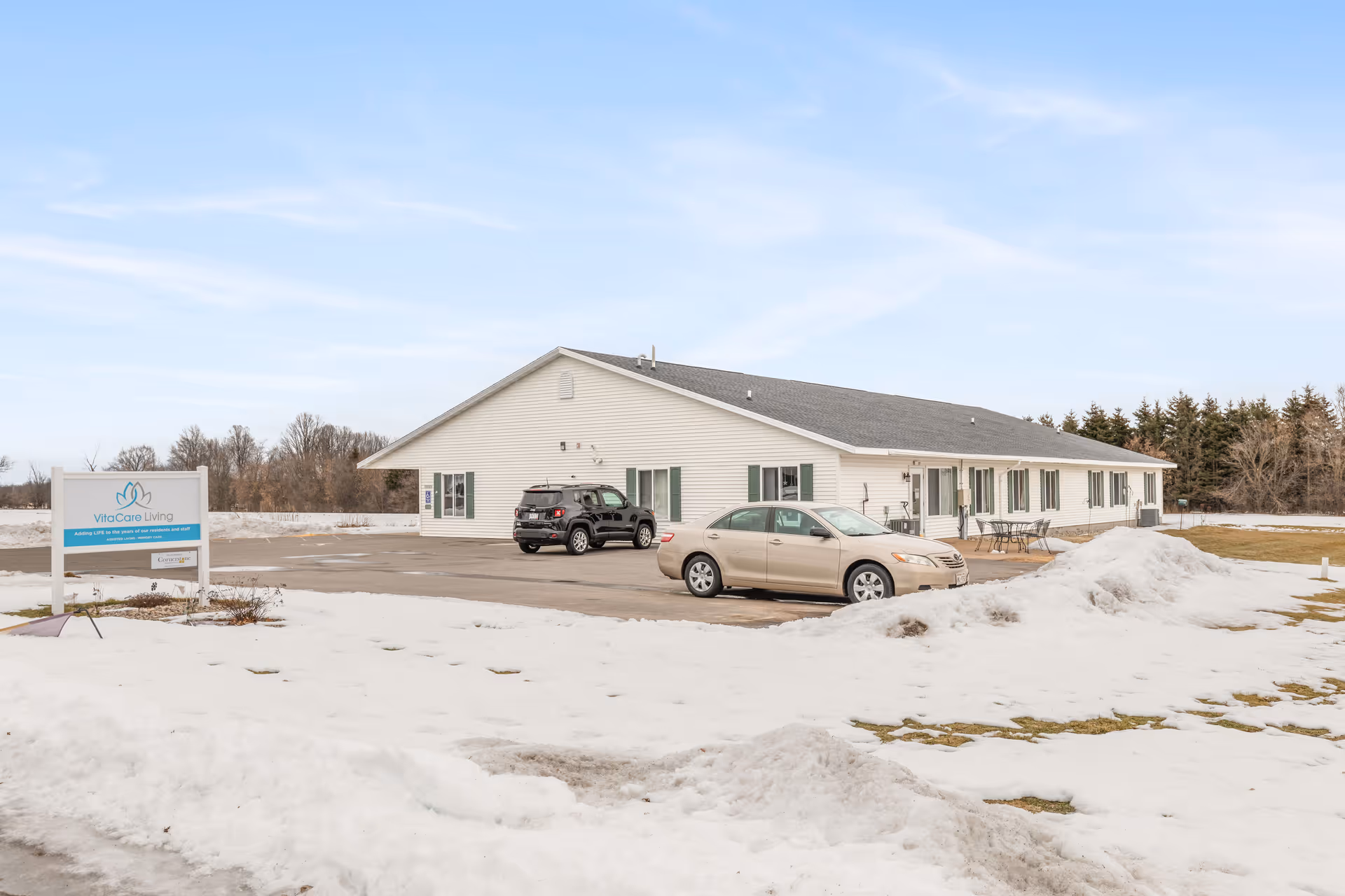 Single-story white senior living building with parked cars, a VitaCare Living sign, and snow-covered grounds.