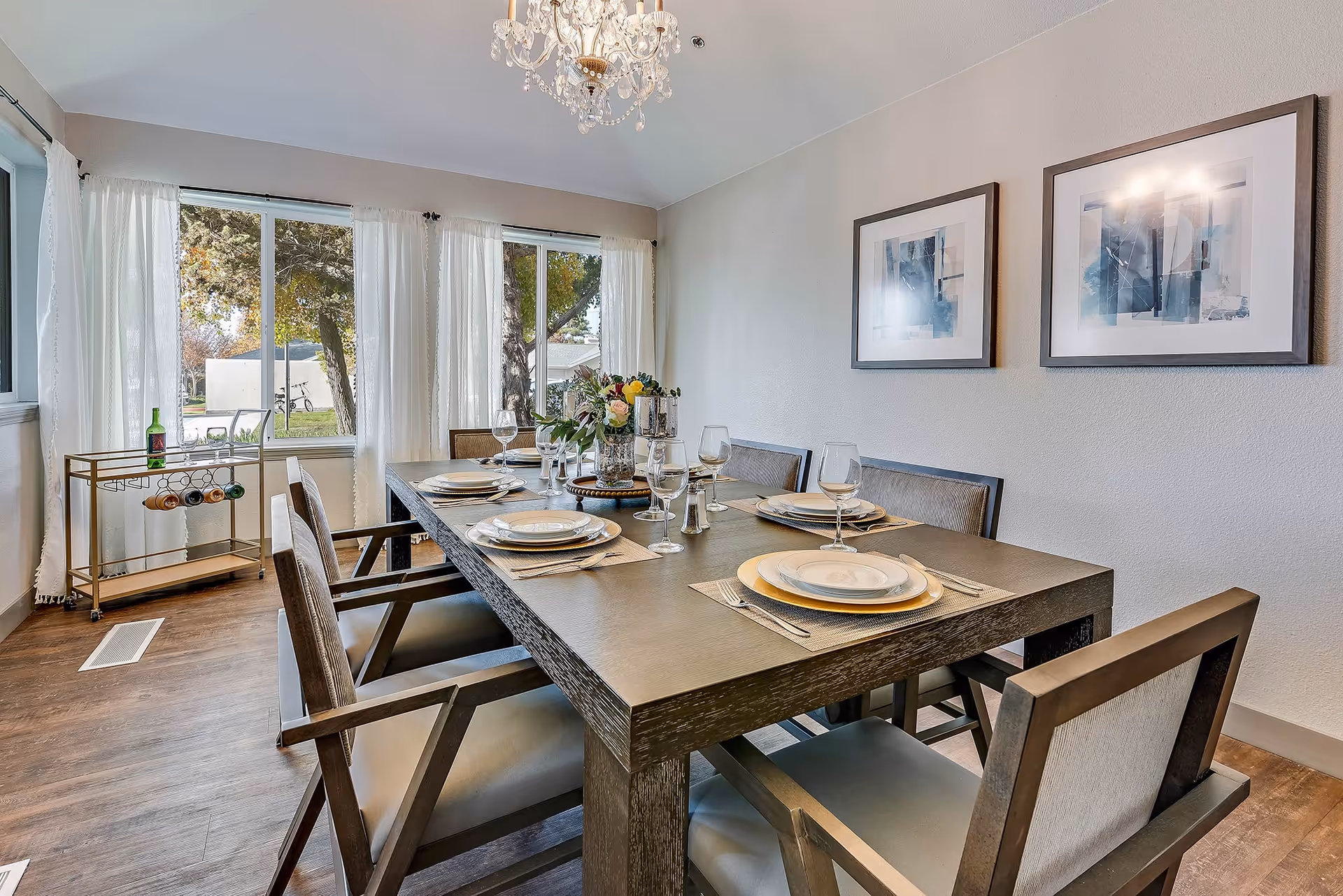 Bright dining room with a large wooden table set for six, modern chairs, a chandelier, and windows with sheer curtains.