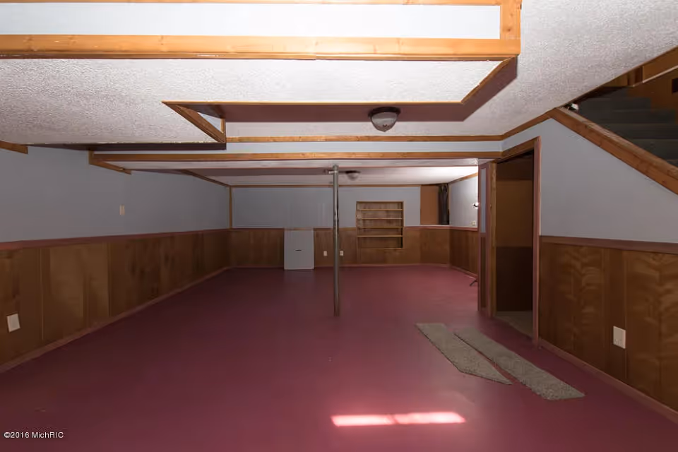 Empty basement room with maroon floor, wood-paneled lower walls, gray upper walls, and a white textured ceiling with wooden beams. There is a metal support pole in the center, a small built-in wooden shelf on the far wall, and stairs leading up on the right side.