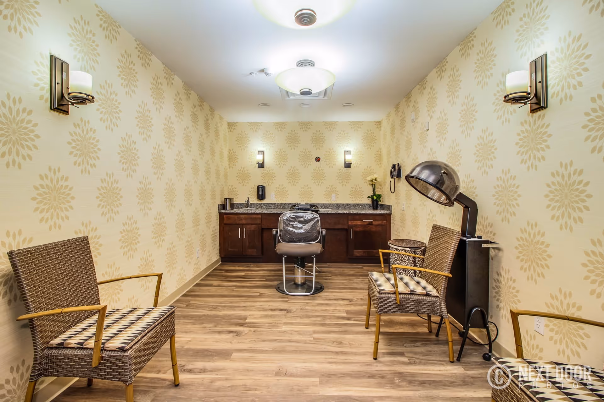 A small salon room with two wicker chairs with patterned cushions, a salon chair in front of a countertop with cabinets, a hair dryer hood on a stand, and floral patterned wallpaper on the walls.