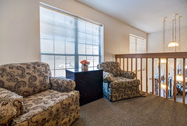 A cozy sitting area with two patterned armchairs and a small black chest of drawers between them, placed near large windows with blinds. The area overlooks a lower level with hanging pendant lights and a railing.