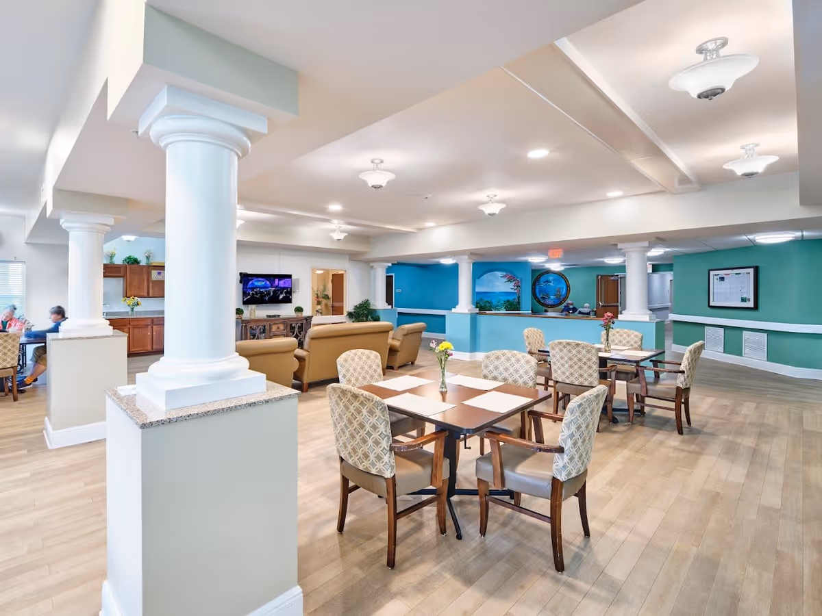 A bright and spacious common area in a senior living facility featuring several tables with chairs arranged for dining or socializing. The room has light wood flooring, white columns, and teal accent walls. In the background, there is a seating area with beige sofas and a wall-mounted television. The space is well-lit with ceiling lights and decorated with small vases of flowers on the tables.