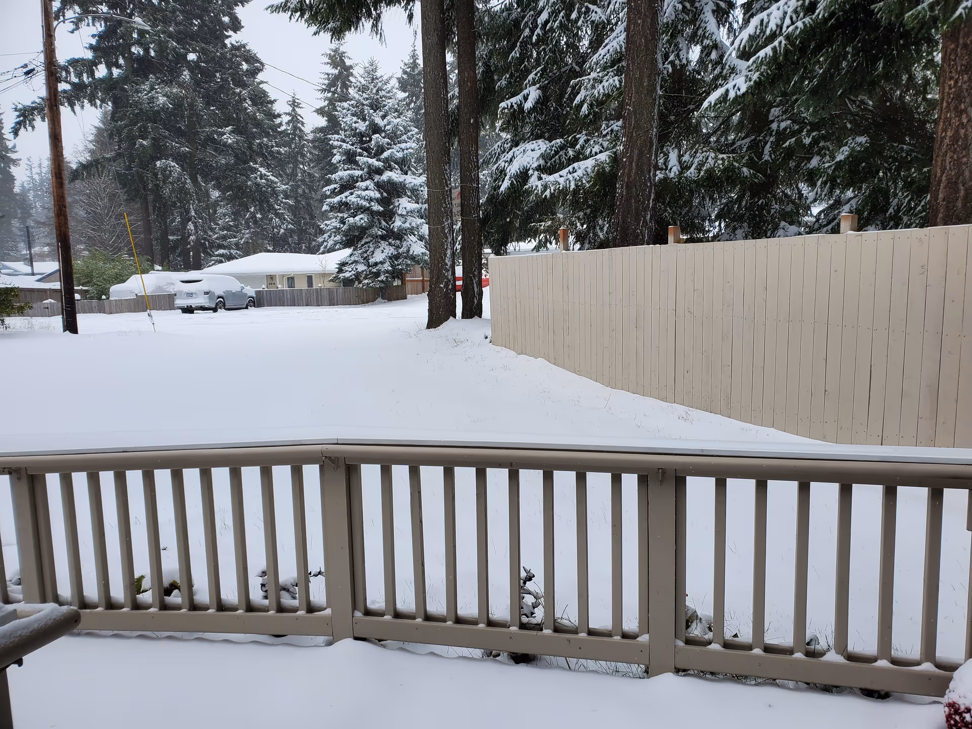 Snow-covered backyard with a beige wooden fence and railing in the foreground, surrounded by tall evergreen trees also covered in snow. A snow-covered car and a house are visible in the background.