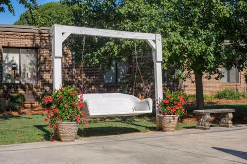 Outdoor courtyard with white hanging swing, flower pots, and a stone bench beside a brick building.