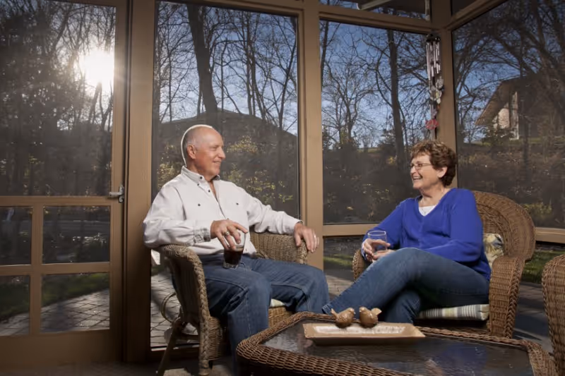 An elderly man and woman sitting in wicker chairs on a screened-in porch, enjoying drinks and smiling at each other. The sun is shining through the screened windows, and there are trees visible outside.