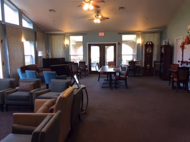 A spacious senior living common area with multiple seating arrangements including armchairs and tables. The room has large windows letting in natural light, ceiling fans with lights, a piano, and a grandfather clock. There are double glass doors leading to an outdoor patio area.