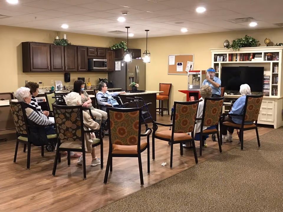 A group of elderly people seated in a semi-circle on patterned chairs in a common room with a kitchen area in the background. A staff member wearing a face shield is standing near a popcorn machine, engaging with the group. The room has wooden flooring, beige walls, and a large white entertainment center with a TV and bookshelves.